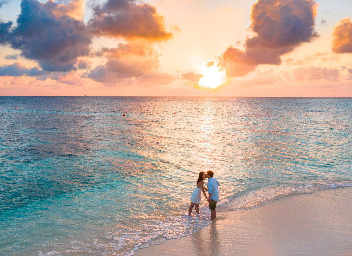 A couple standing in shallow turquoise ocean water at sunset on a sandy beach, with glowing orange clouds and reflected light on the water.