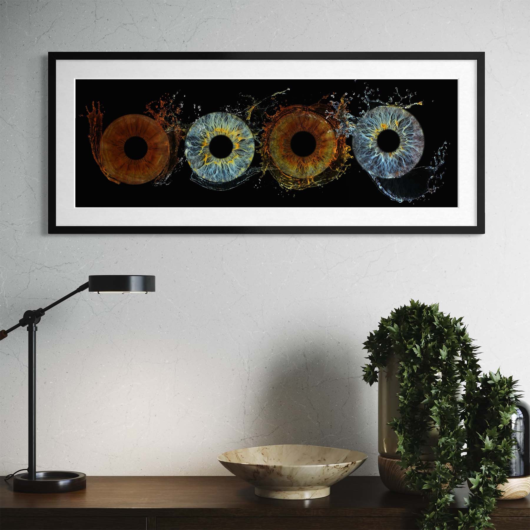 Framed artwork of colourful eye  designs on a wall above a desk with a lamp and plant.