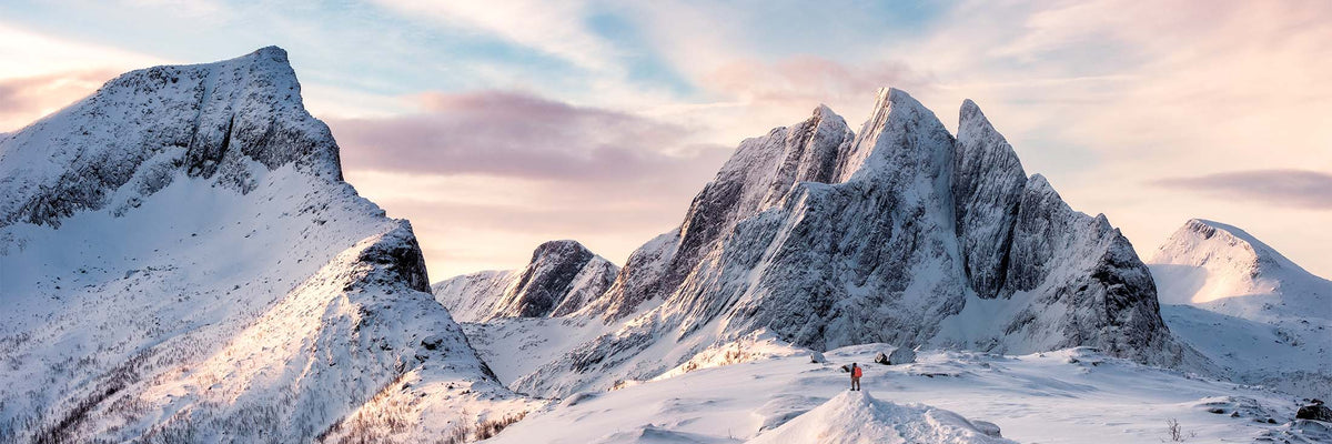 A dramatic snowy mountain landscape with jagged peaks, snow-covered slopes, and a pale sky tinged with pink and blue light. A tiny person appears near the lower center-right on the snow, emphasizing the vastness of the scene.