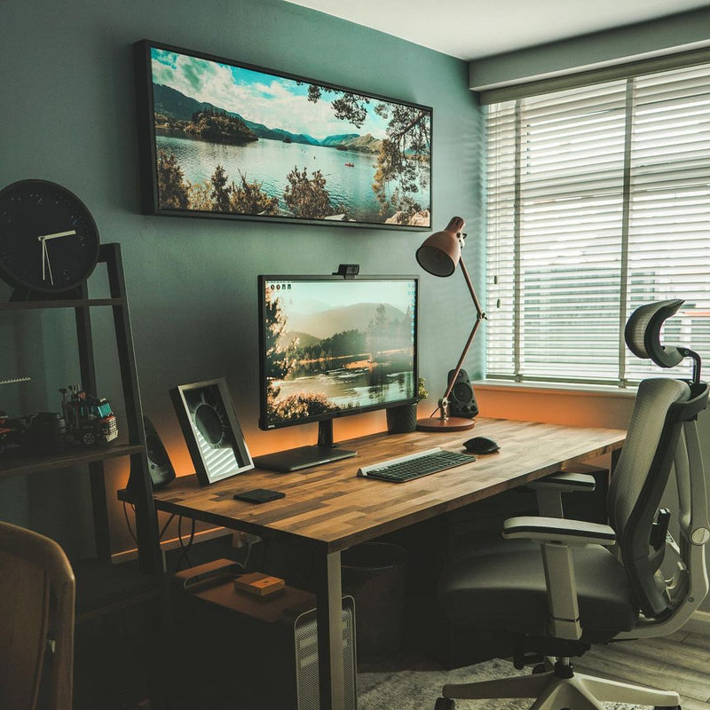 A modern home office setup featuring a wooden desk with a dual-monitor setup, a black ergonomic office chair, a wall-mounted floating framed canvas print, and a dark teal wall.