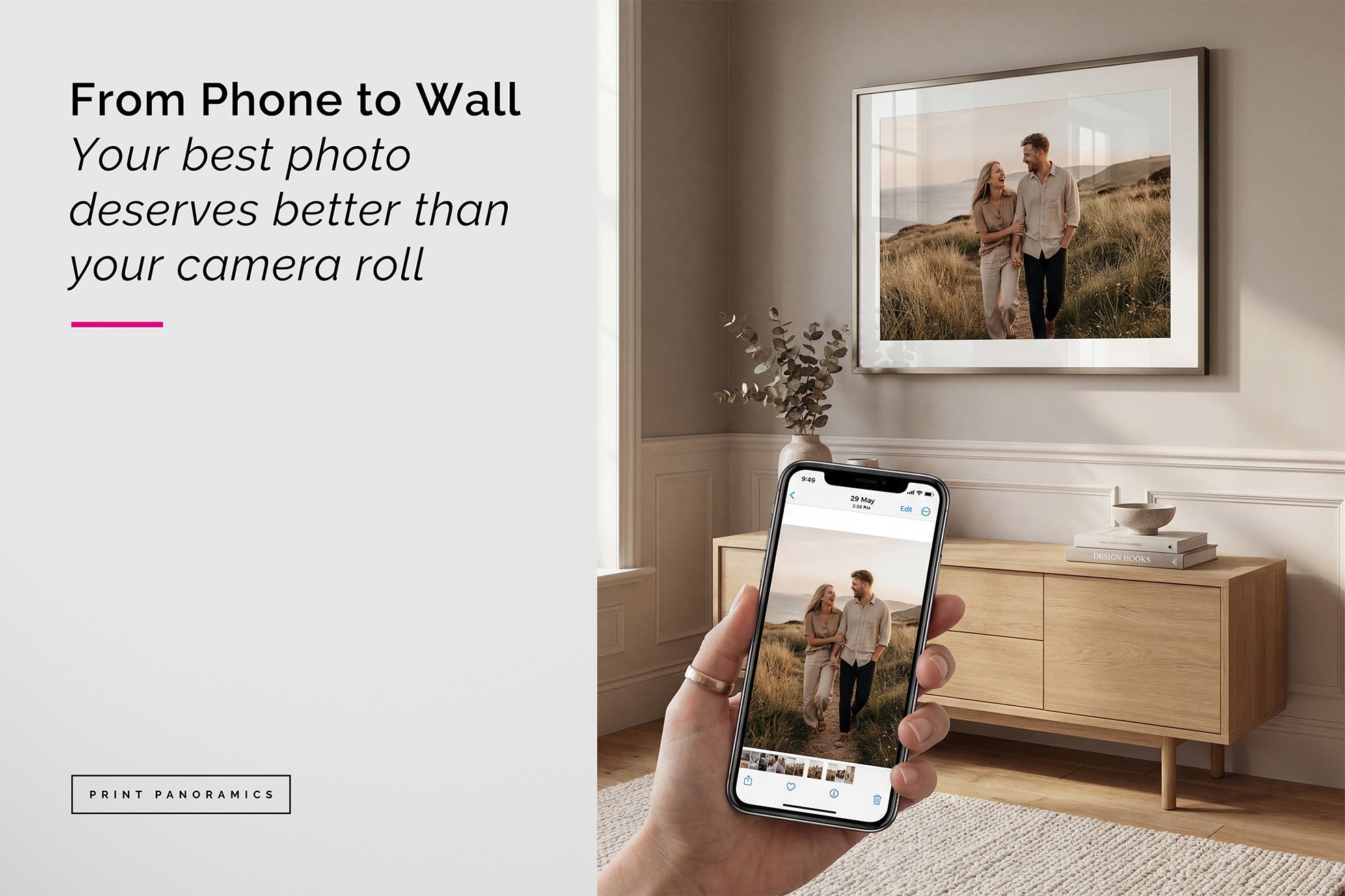 Hand holding a smartphone with a photo displayed, showing a framed mounted photo print of a couple in a field on a wall.