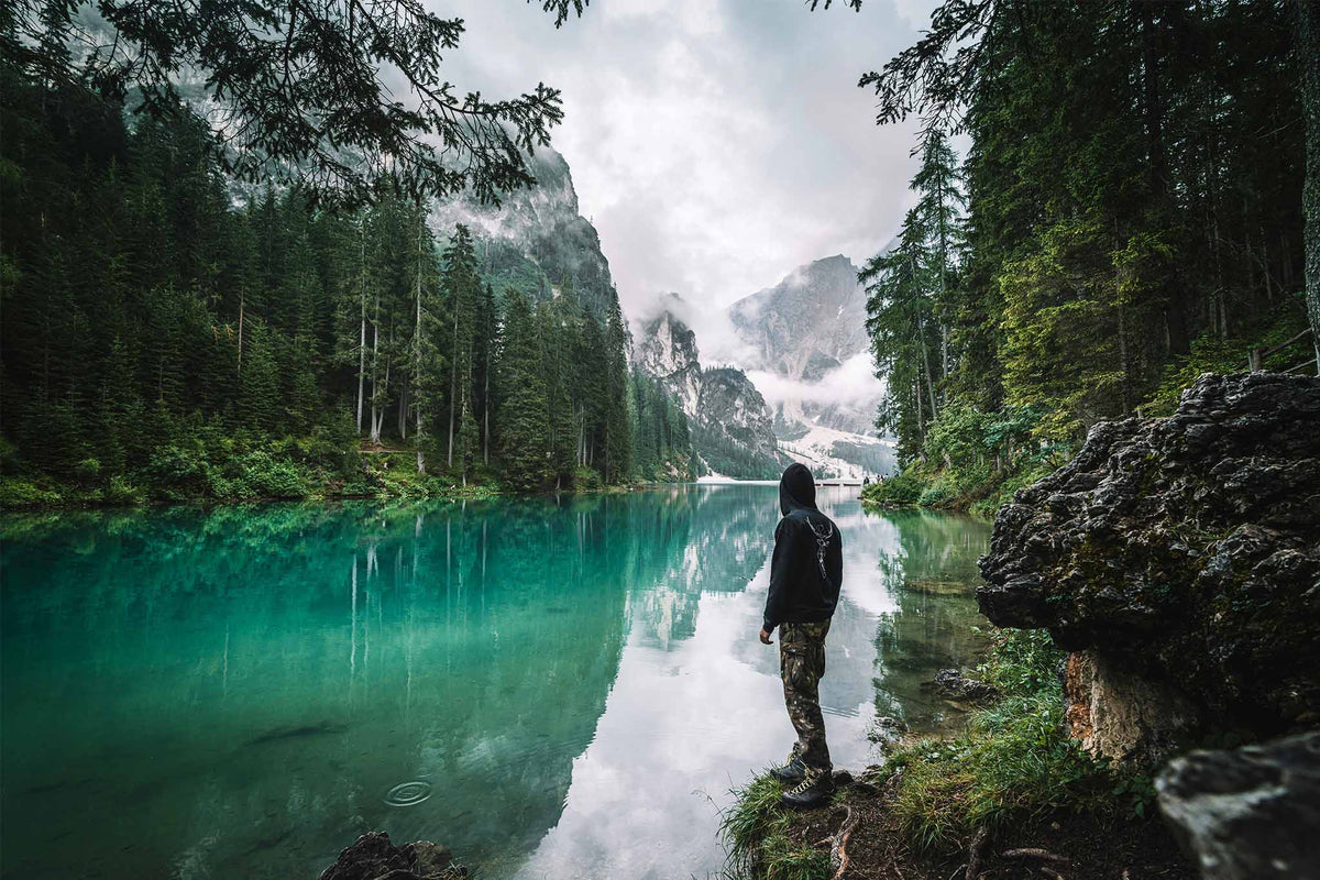 A person in a black hoodie and camouflage trousers stands on the edge of a turquoise alpine lake, looking out at a misty mountain valley framed by a dense evergreen forest.