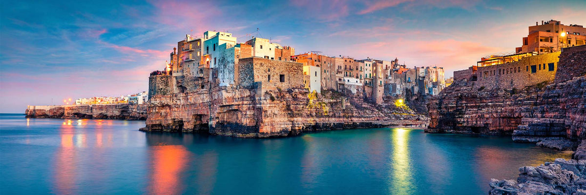 A scenic coastal town perched on rocky cliffs above calm blue water, with warm glowing buildings under a colorful sunset sky.