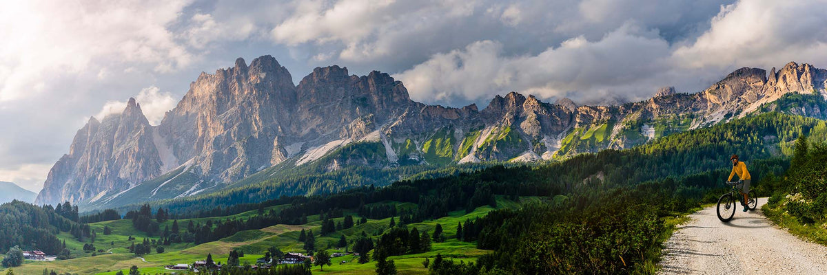 A scenic mountain landscape with jagged rocky peaks, green valleys, and a gravel path where a cyclist rides in the distance under a dramatic cloudy sky.