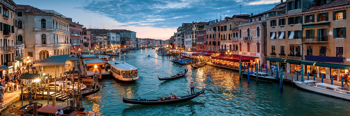 A scenic Venice canal view at dusk with gondolas on the water, illuminated buildings lining the canal, and warm lights reflecting on the blue-green water.