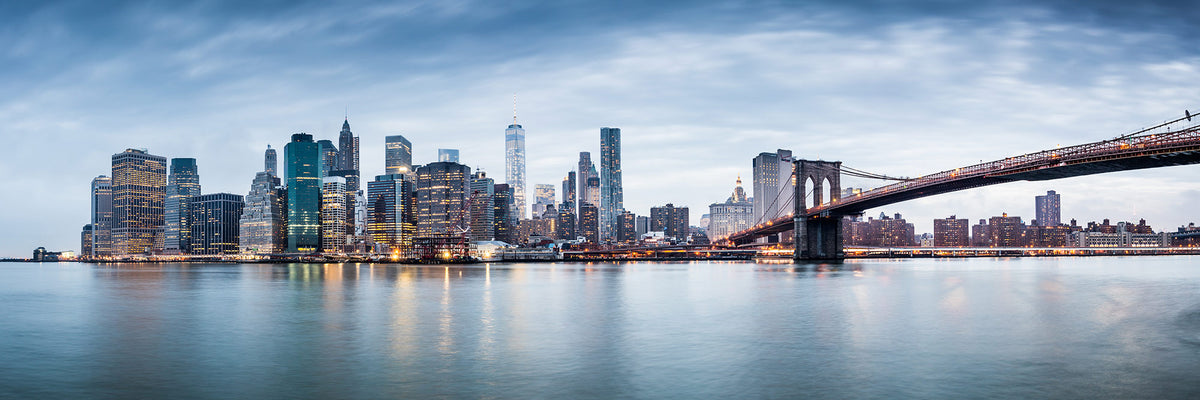Panoramic view of the New York City skyline across calm water, with the Brooklyn Bridge extending from the right side under a cloudy blue sky.