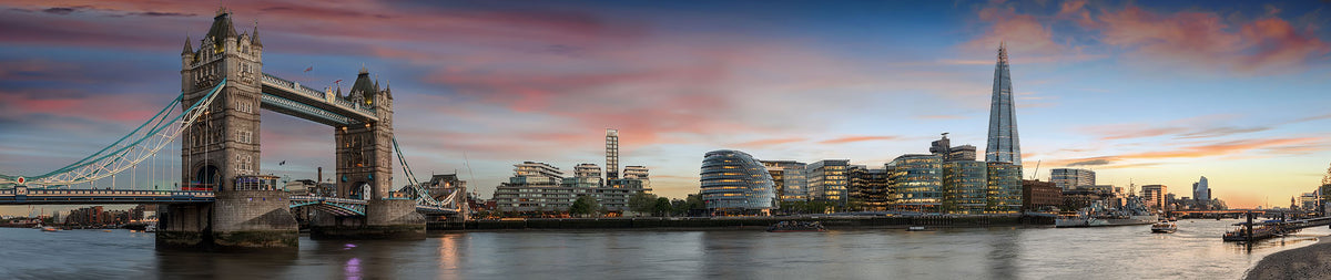 Panoramic view of the London skyline at sunset, featuring Tower Bridge on the left and the Shard on the right, with the Thames in the foreground and a colourful sky in pink, orange, and blue tones.