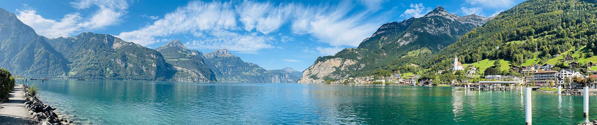 A scenic panoramic landscape of a mountain fjord with clear turquoise water, steep green and rocky peaks, a small village with white buildings, and a bright blue sky with wispy clouds.